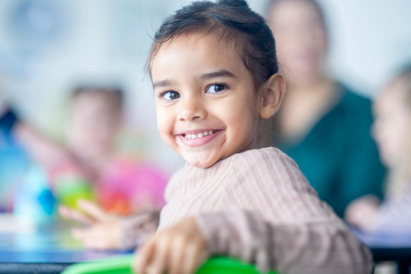 Asian girl child with a warm smile in a bright classroom, surrounded by adults and colorful objects. A joyful moment, perfect for themes of education, family, and positivity in diverse settings.