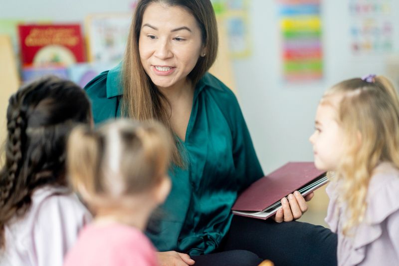 A warm classroom moment where an Asian teacher interacts with young students, offering guidance and support during learning. Calm, friendly atmosphere emphasizing education, care, and engagement.