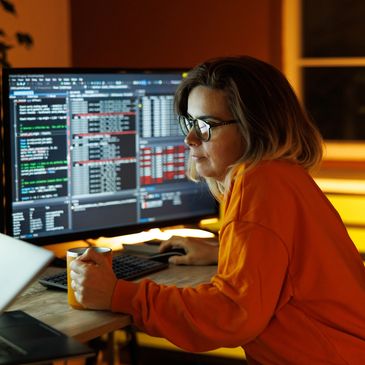 Woman in glasses working late at a computer with code on screen and holding a mug.