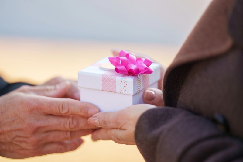 Close-up view of an elderly man handing a small gift box to a woman outdoors during daytime. The focus is on their hands as the gift is being given and received, with the box wrapped and decorated with a ribbon. The image represents gift giving, surprise, and appreciation between two senior adults in an outdoor park setting, with selective focus and copy space.
