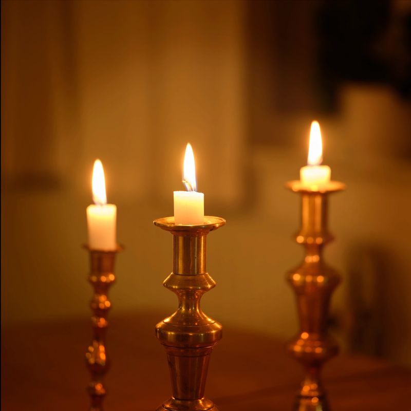 A serene interior scene featuring four golden brass candlesticks with tall white candles set on a wooden table. In the softly lit background, a piano rests beside a vase of flowers and a leafy plant, creating a warm, reverent atmosphere perfect for seasonal, ceremonial, or reflective themes.