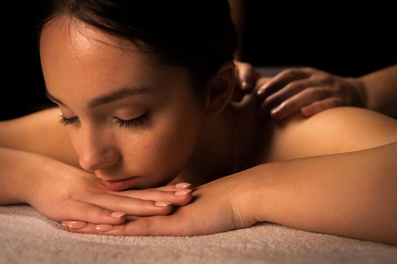 A young woman lies face down, receiving a gentle massage. Her hands rest on a soft surface, eyes closed, exuding calm. The serene spa setting emphasizes relaxation and self-care, highlighting beauty and therapeutic benefits.