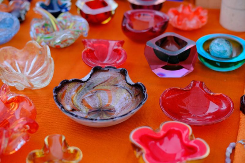 Colorful handmade glass bowls and decorative objects displayed on a market table. The vibrant shapes, textures, and translucent colors highlight artisanal craftsmanship, creativity, and contemporary design, evoking traditional glassmaking and artistic expression. No people visible.