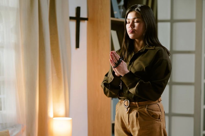 Asian woman standing with eyes closed holding rosary beads and praying in front of wooden cross in home interior, hands clasped together in front of chest