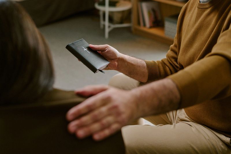 Middle aged Caucasian man comforting by placing hand on shoulder while holding closed Bible, engaging in supportive gesture indoors, focus on hands and book