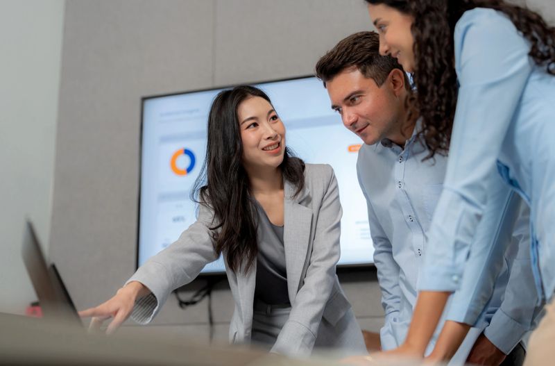Professional business team analyzing annual sales data on a monitor. Confident woman pointing at financial charts and explaining market trends to colleagues during a strategic meeting in a modern corporate office.