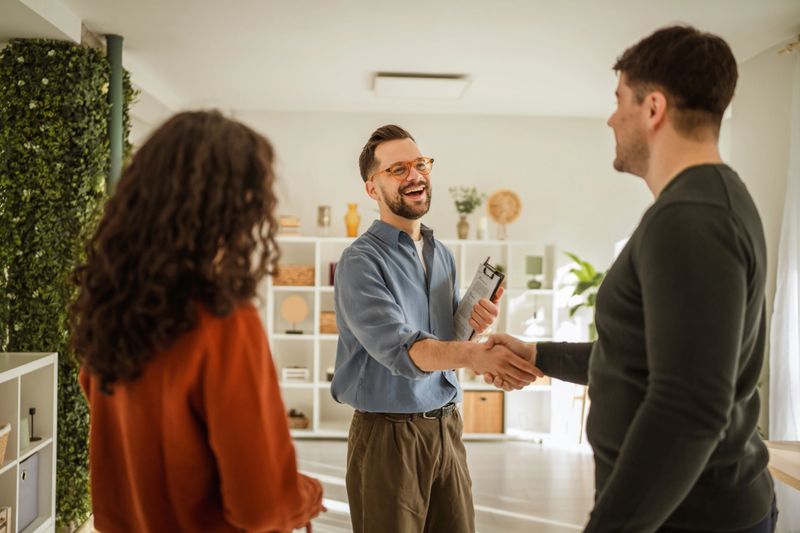 Real estate agent smiling while shaking hands with a new homeowner, signifying a successful agreement and closing a property deal for the couple in a modern home