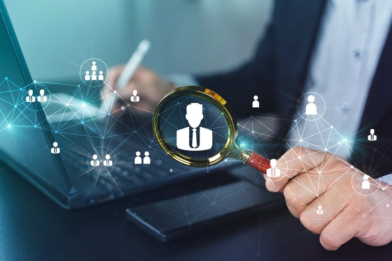 Business professional using magnifying glass over user icon on laptop, showing network connections for HR recruitment, customer search, identity verification, and data security analytics.