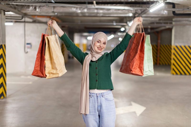A smiling woman wearing a hijab holds shopping bags in a parking garage, celebrating her purchases