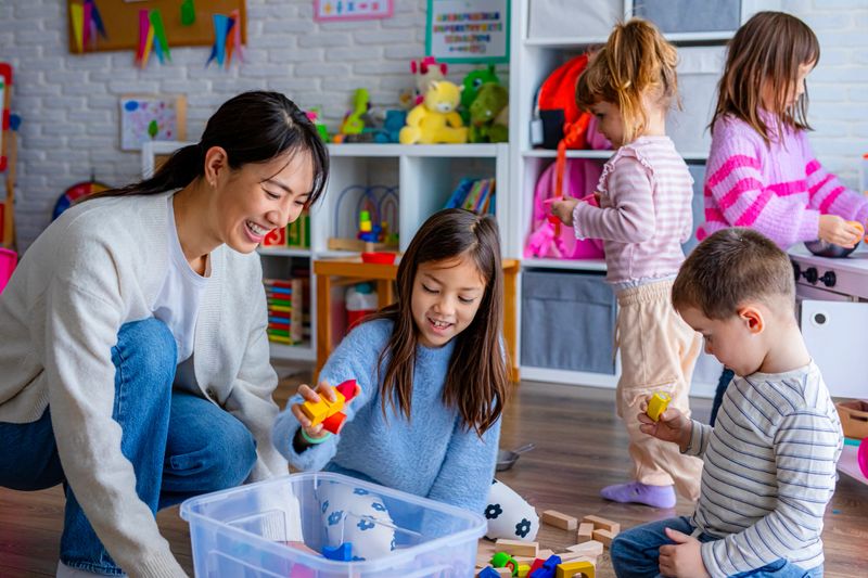 A preschool teacher guides children as they collect and organize toys together after a group play activity in the classroom