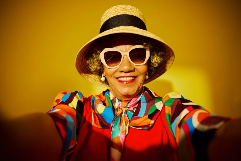 Senior woman smiling, wearing a hat, sunglasses, and colorful clothing, capturing a selfie