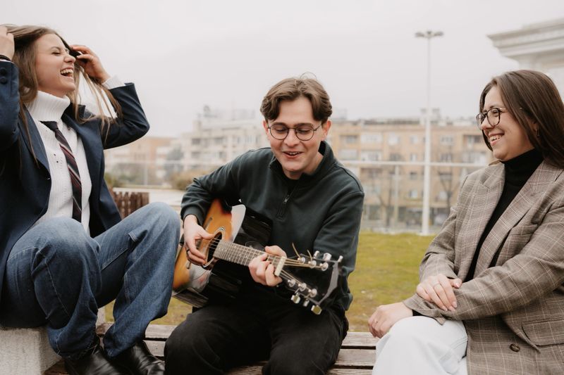 Happy group of young friends laughing and enjoying a spontaneous outdoor music session with acoustic guitar. Concept of joy, youth lifestyle, friendship and positive energy.