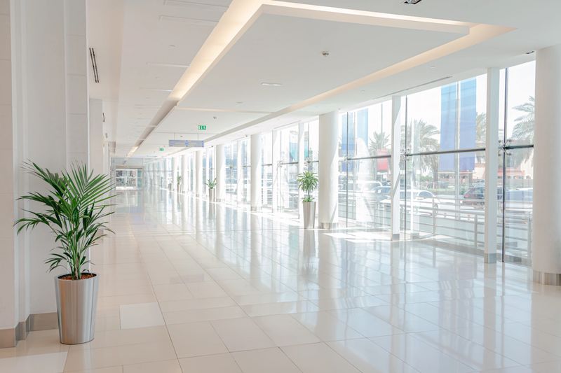 Sunlit empty hallway in a modern shopping mall with clean white tile floor, tall pillars, glass wall, indoor potted palms in metal planters, reflections and long perspective creating a minimalist architectural background with ample copy space for retail, airport, hospital or office concepts