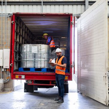 Workers loading metal drums into a truck in a warehouse.