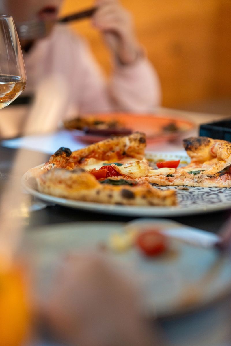 Pizza slices served on a patterned plate at an Italian restaurant dining table. The focus is on the deliciously baked crust and toppings. Nearby, diners are enjoying their meal, creating a lively dining atmosphere.