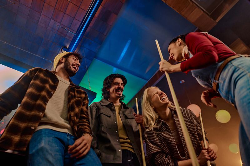 A group of friends share a fun moment playing billiards in a cozy pub, laughing and smiling as they lean over cues. Warm lighting and colorful background create a lively, social atmosphere.