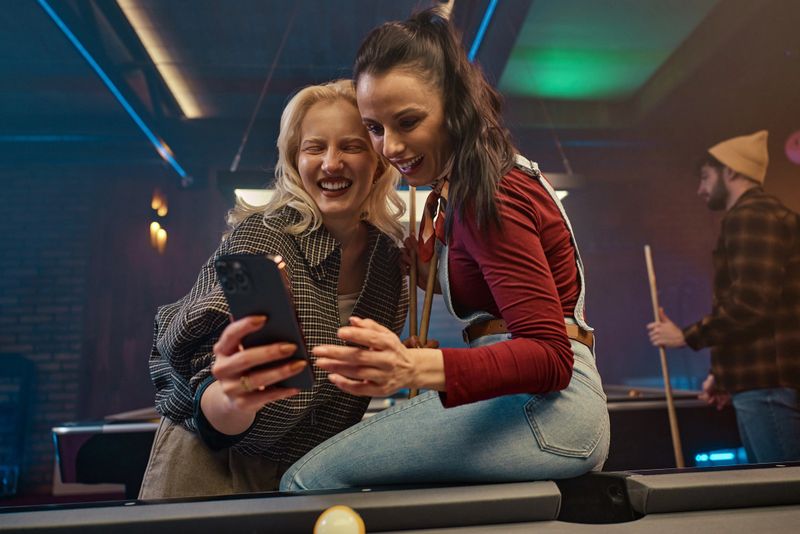 Two women share a laugh while taking a selfie at a pool table, with a man in the background. The scene captures friendship, casual nightlife, and a fun, relaxed mood in a pub.