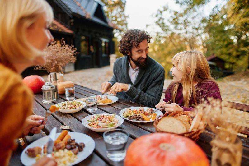 Happy family eating lunch together at a rustic wooden table in the backyard during fall
