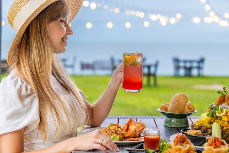 Woman in summer hat enjoying seafood meal with tropical drink at outdoor restaurant near beach. Relaxation, vacation, dining, and leisure concept.