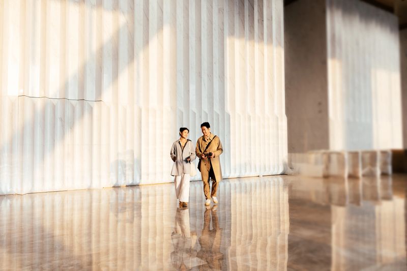 Wide view of two confident Asian businesspeople  talking and walking enter a large, light filled interior space together after working after working.