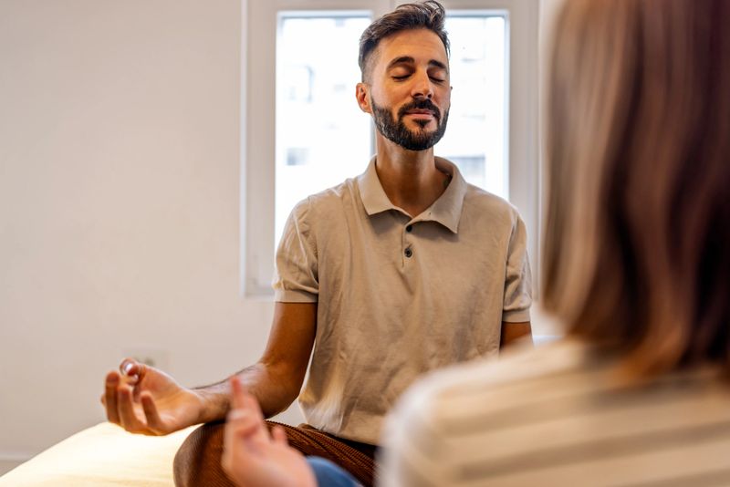 A man sits calmly with eyes closed, practicing meditation during a wellness or therapy session. A listening therapist sits nearby, creating a serene, supportive atmosphere with soft window light.