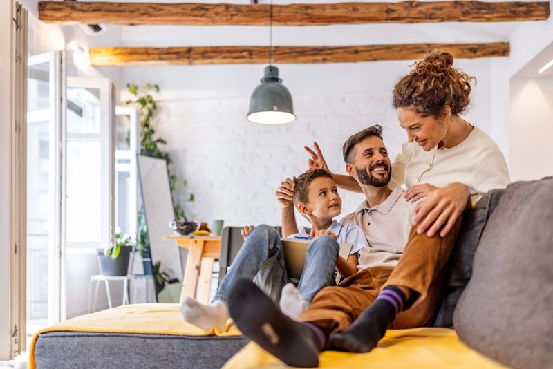 A cheerful family moment at home as parents and their child sit together on a sofa, sharing smiles and playfulness in a bright living room with wooden beams and plants.