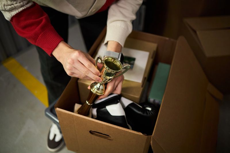 Young adult Caucasian man packing cardboard box with personal belongings, holding small trophy in one hand, standing indoors, only upper body and hands visible