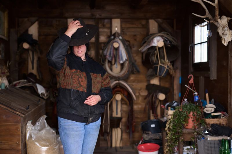 A cowgirl in a dark jacket and hat stands in a wooden tack room filled with saddles, ropes, and plants. She touches her hat, exuding confidence in a traditional country setting.