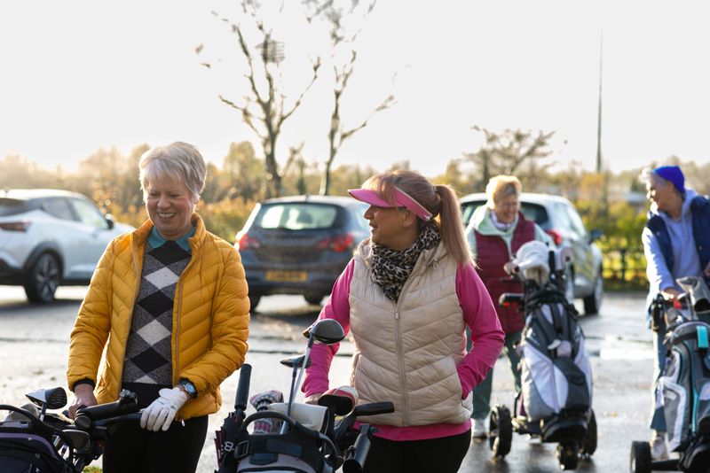 A wide shot of a group of mature women walking toward the golf course with their golf trolleys, smiling and chatting in a car park before play. The image highlights friendship, active aging, and the social enjoyment of golf among older women.Videos are available similar to this scenario.