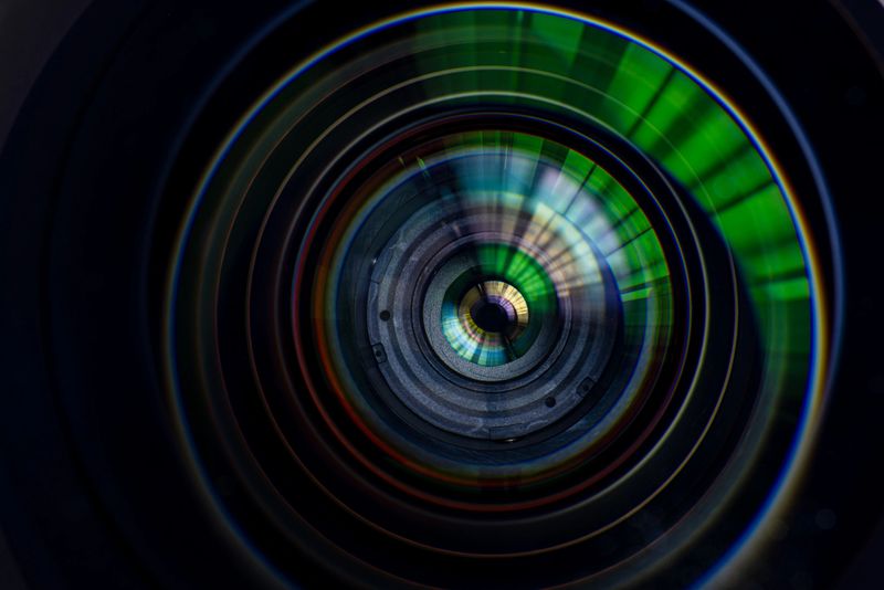 A detailed, front-facing macro shot of a 16-35mm wide-angle camera lens, highlighting the intricate multi-coated glass elements, internal optics, and the reflection of light within the barrel. The image showcases the precision engineering of professional photography equipment with a dark, cinematic aesthetic.