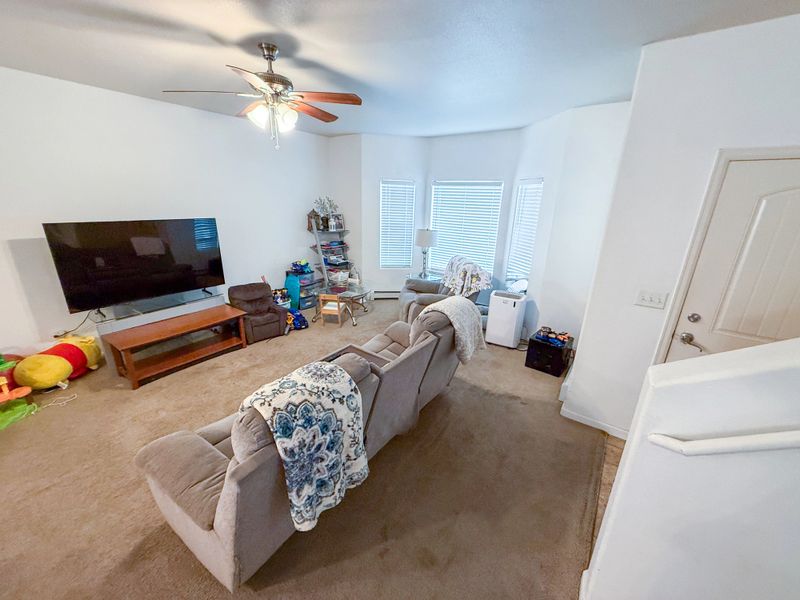Wide-angle interior shot of a clean, affordable housing living room featuring carpeted floors, neutral recliner seating, a television, a ceiling fan, and natural daylight through multiple windows. The image represents low-income housing, rental housing interiors, and everyday residential living in a modest, maintained home environment.