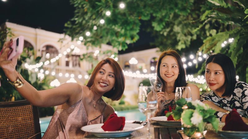 asian women taking selfies during an outdoor dinner, reflecting digital social behavior and lifestyle storytelling. the scene supports themes of social media culture, modern friendship, experiential dining, and content creation within premium leisure environments.