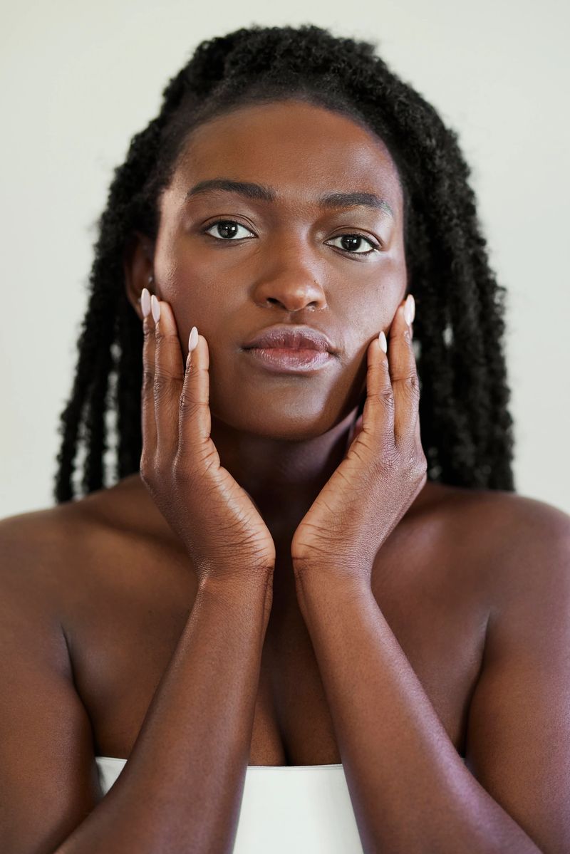 Vertical beauty portrait of a woman with dark skin and textured hair twists touching her cheeks with both hands.
