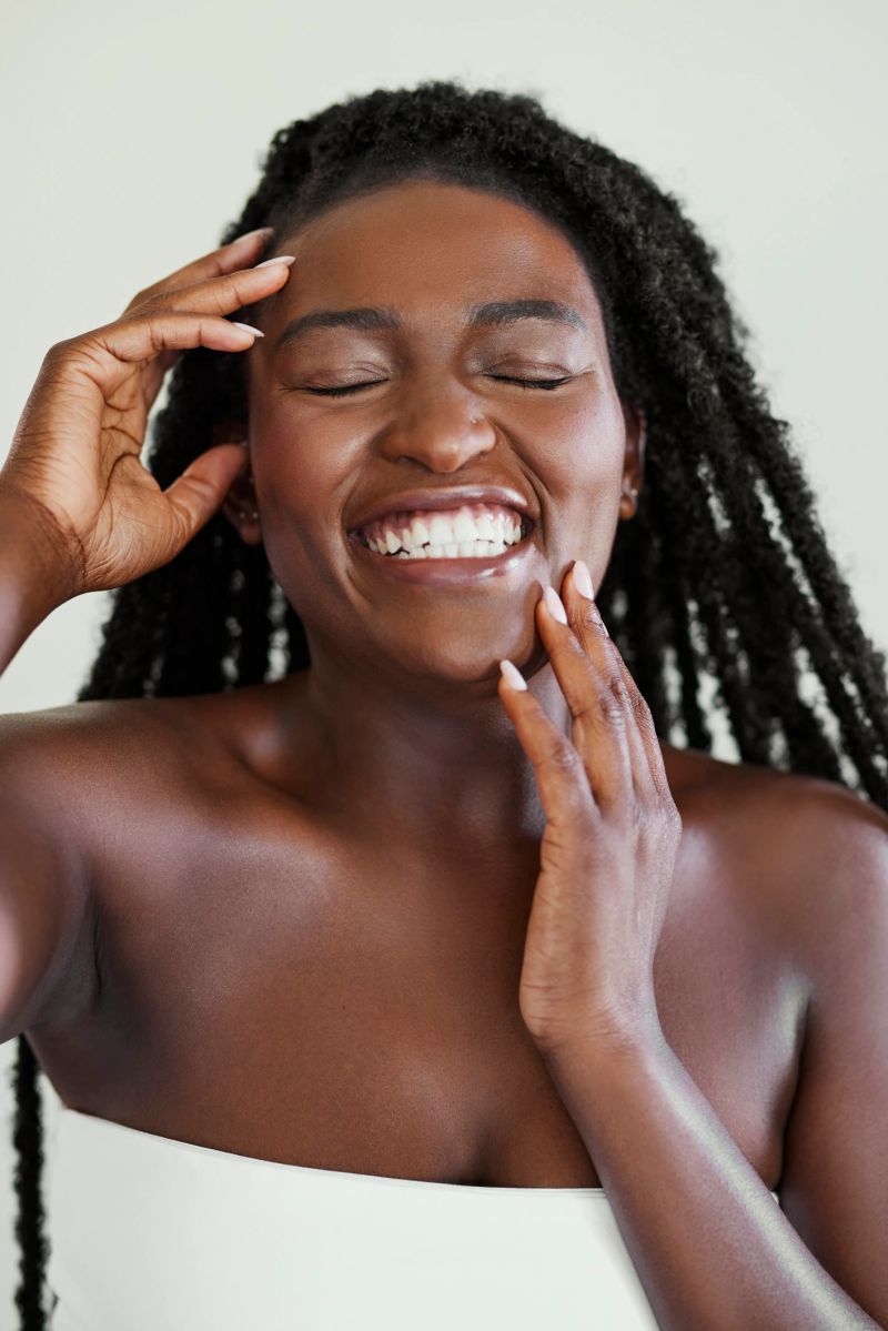 Vertical beauty portrait of a joyful Black woman with long twists laughing heartily while touching her glowing skin.