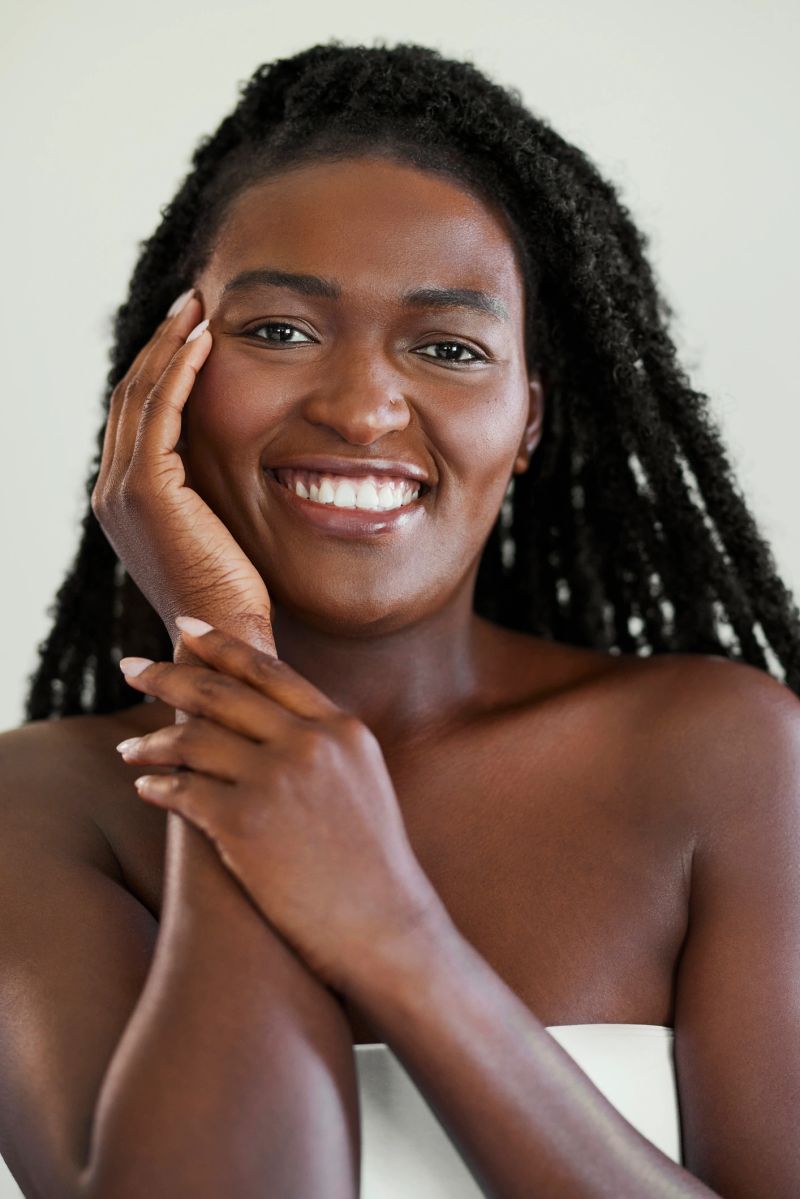 Vertical beauty portrait of a happy Black woman with glowing skin and long hair twists smiling while touching her face.