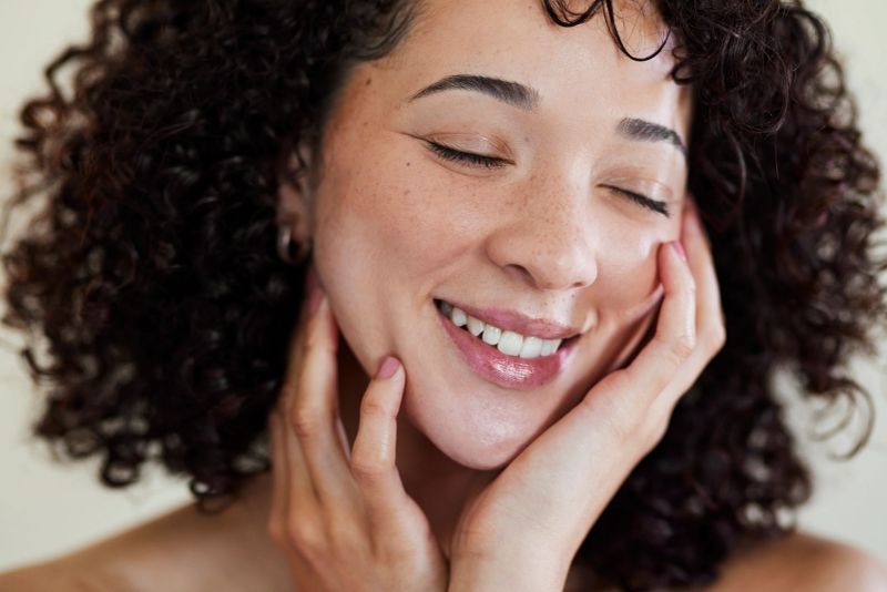 A horizontal close-up of a joyful young woman with curly hair and freckles smiling while touching her radiant skin.