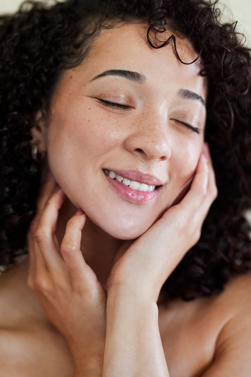 A vertical close-up of a peaceful young woman with curly hair and freckles, smiling with her eyes closed while gently touching her radiant face.