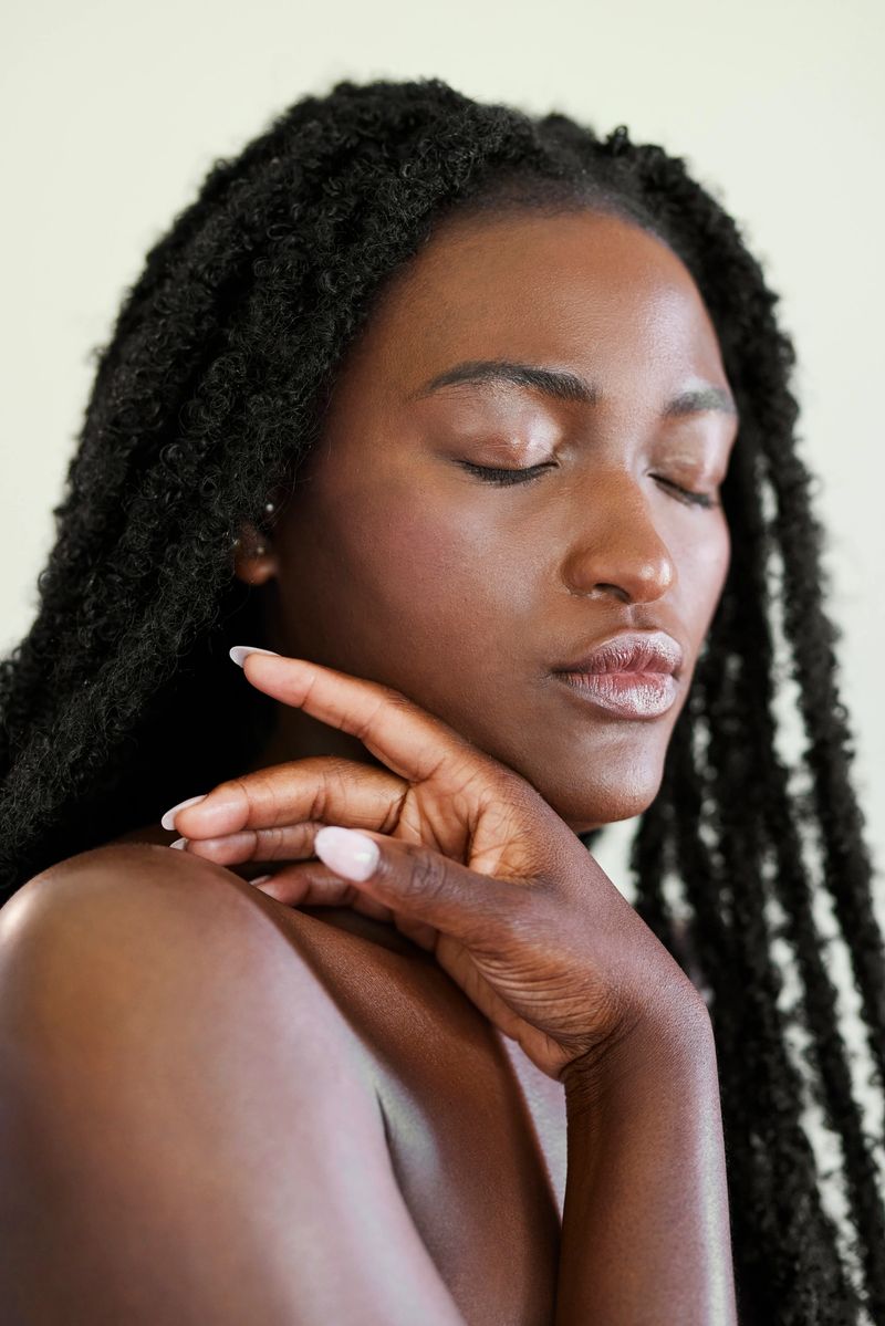 A vertical close-up of a serene African American woman with eyes closed, gently touching her radiant skin to showcase natural beauty.