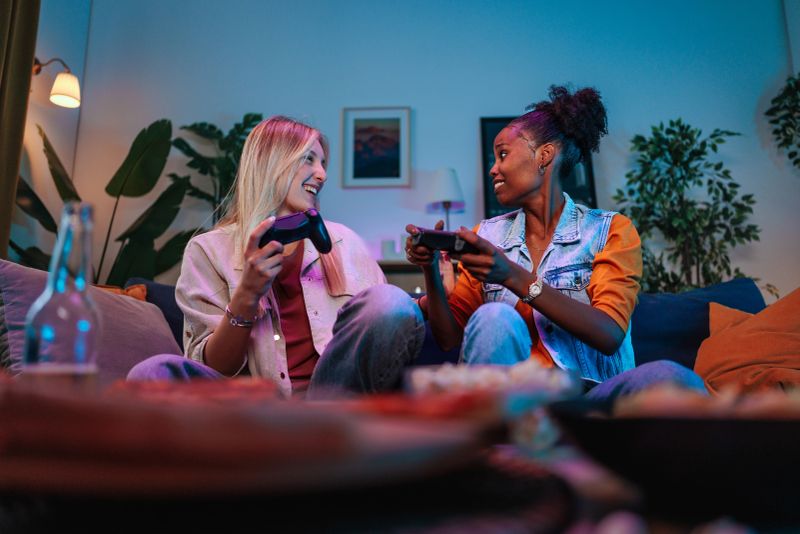 Two women share a lively game night on a cozy living room couch, holding game controllers and laughing under blue and purple ambient lighting. A bottle and snacks sit nearby.