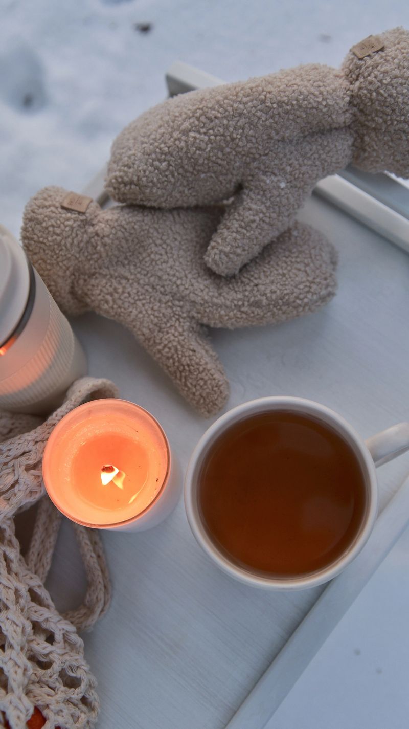 A comforting arrangement of a steaming mug of tea, a lit candle, and soft mittens sits on a tray amidst a snowy landscape, evoking warmth and tranquility