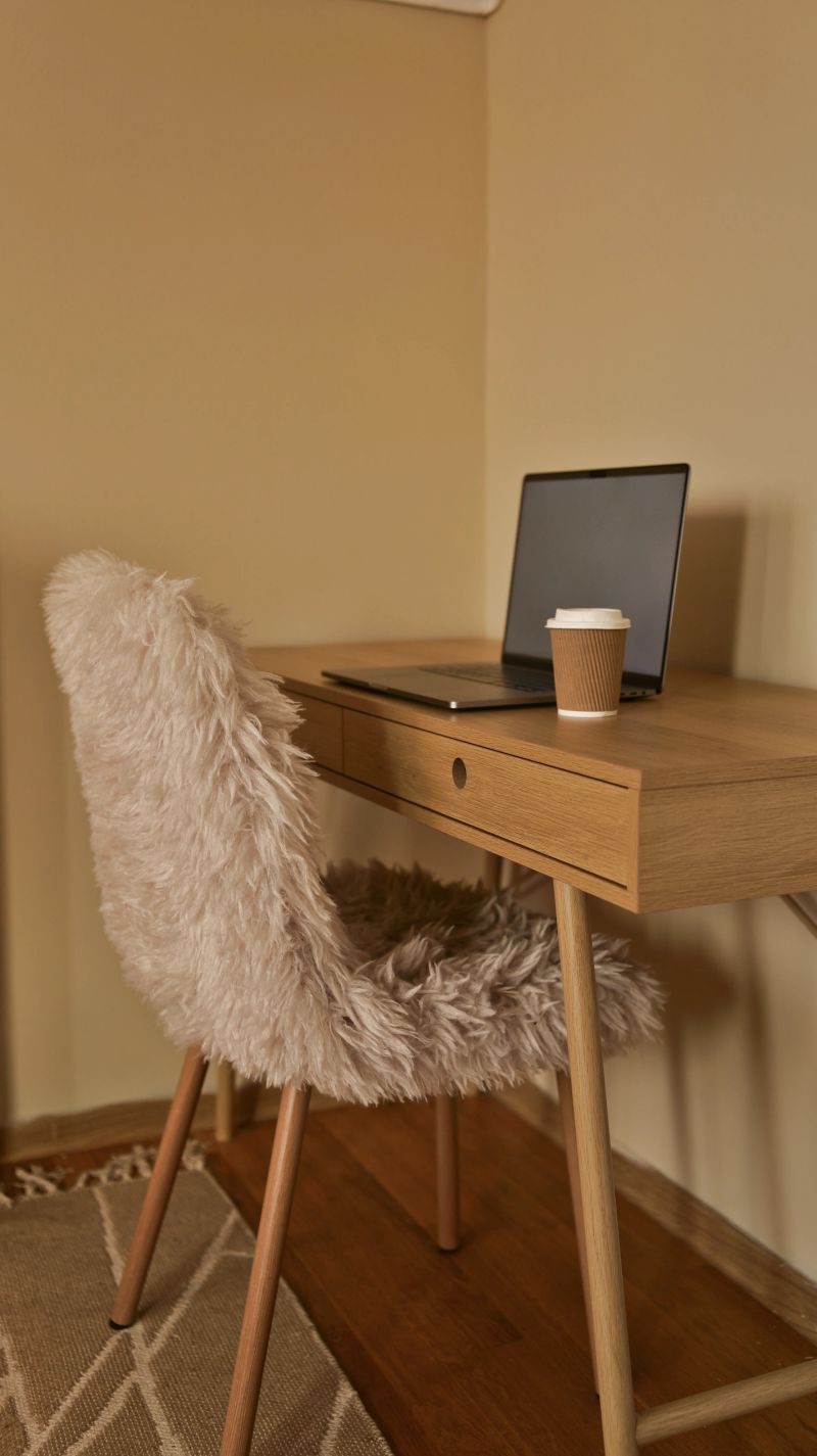 A modern wooden desk with a laptop and a disposable coffee cup is paired with a plush, fluffy chair for a comfortable workspace