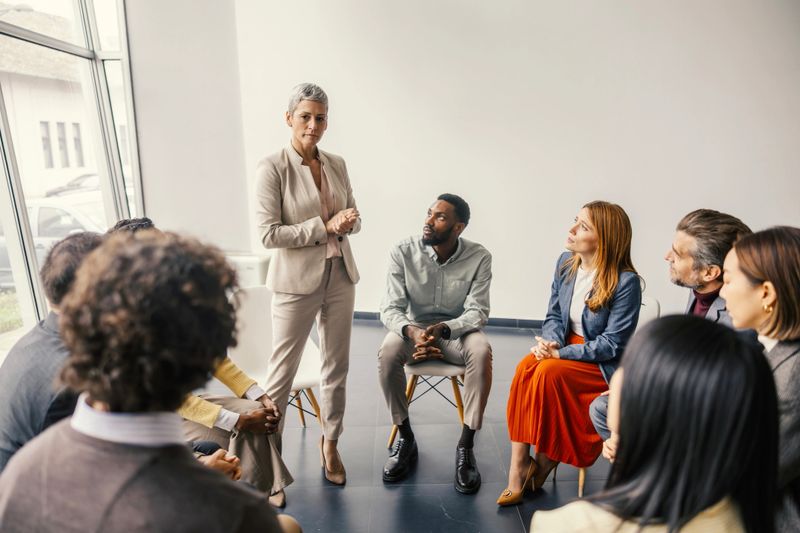 Mature adult business leader standing, addressing a diverse group of professionals sitting in a circle during a corporate training session or workshop, fostering collaboration and communication
