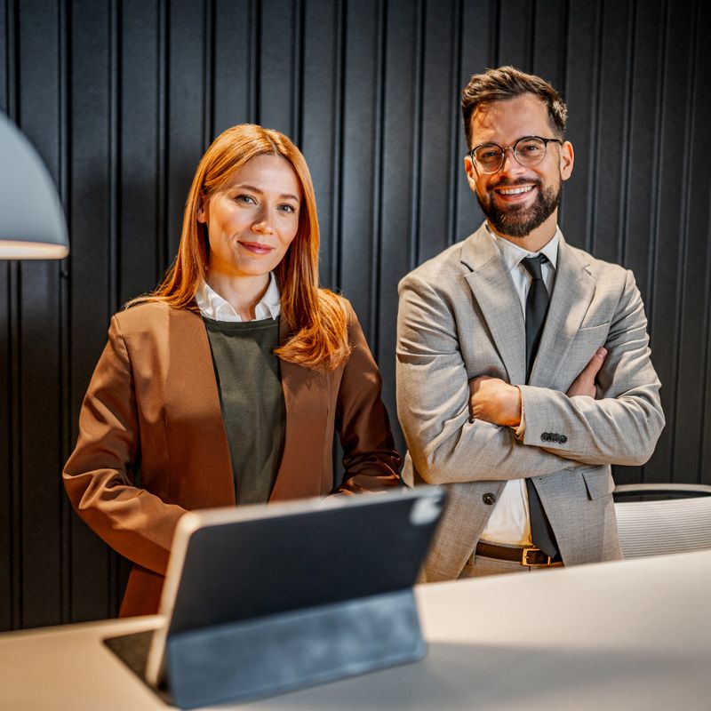 Two smiling business professionals, a man and a woman, standing together in a modern office lobby, ready to assist or greet