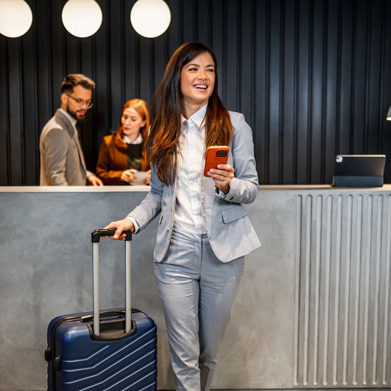 Professional businesswoman arriving at hotel lobby with suitcase and smartphone, smiling while checking in at reception desk