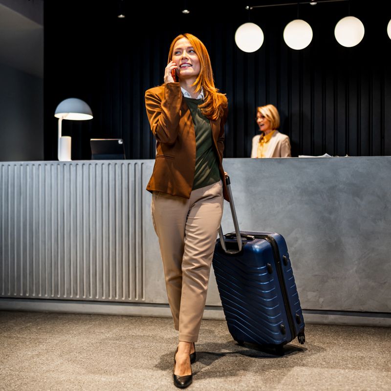 Young adult woman pulling suitcase and talking on smartphone, smiling while arriving at a modern hotel lobby reception