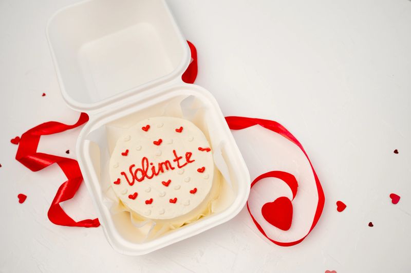 Bento cake. Decorated with red hearts, Volim te (I love you) lettering. Sitting in a white takeaway box. Surrounded by red ribbon, small heart confetti. Loving celebration, romantic gift