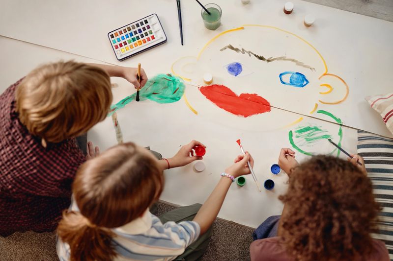 Group of Generation Alpha children painting large face illustration on paper, using watercolor palette and brushes, sitting together on floor, collaborating on creative art project