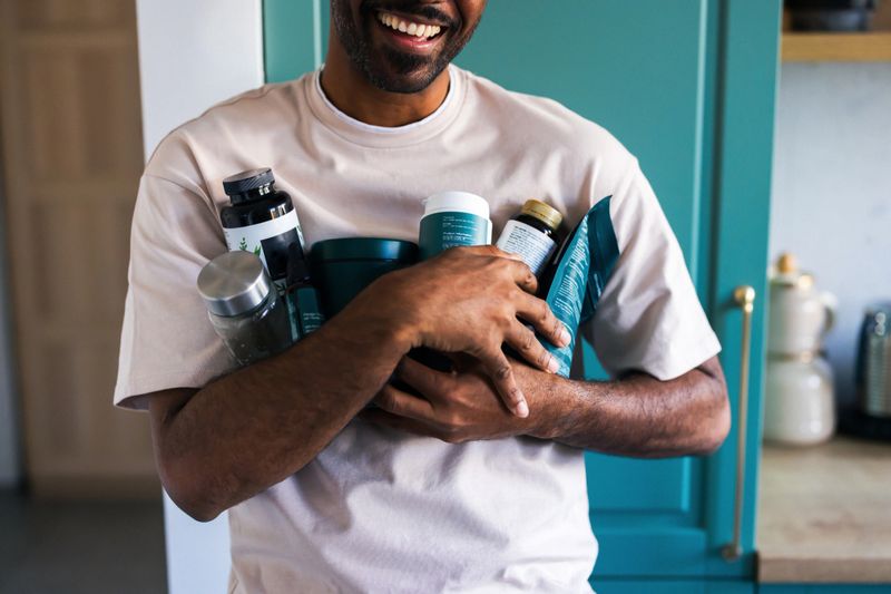 Smiling man holds assorted supplements close to his chest in a bright kitchen.