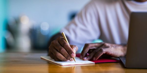 Person writing in a notebook with a pen beside a laptop.