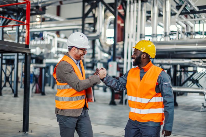 Two smiling multi-ethnic male engineers wearing safety vests and hard hats are shaking hands, celebrating successful partnership inside a modern industrial plant